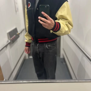 Collegejacka  - Säljer en svart och gul College Jacka med stora emblem på ryggen och bröstet. Jackan har röda och svarta ränder på muddar och krage, samt knappar framtill. Ärmarna är gula och kroppen svart, med cool kontrast och sportig vibe. Finns ett svart märke på armen men syns knappt när man bär den.