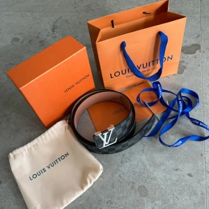 Louis Vuitton rutig skärp i skinn - Säljer ett Louis Vuitton bälte i svart och grått med klassiskt rutigt Damier-mönster och stor LV-logga i silver. Bältet är gjort i exklusivt skinn och kommer med originalpåse, box och påse. Perfekt accessoar för att lyfta din outfit.