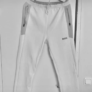 Vita mjukisbyxor från BOSS - Snygga vita mjukisbyxor från BOSS med gråa detaljer och dragkedjeficka på sidan. Byxorna har elastisk midja med snörning och en rak, relaxed passform. Perfekta för chill eller träning och har diskret BOSS-logga framtill. Varan är i ny skick, utan täcken på användning