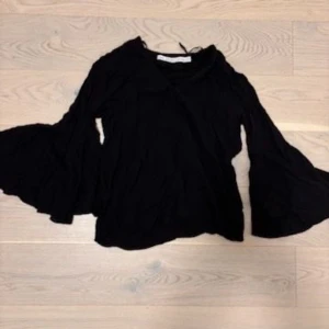 Svart blus med vida ärmar - Svart blus med riktigt vida och fladdriga ärmar som ger en cool och dramatisk look. Tunt och lätt material, perfekt för att sticka ut lite extra. Omlottkänsla vid halsen och avslappnad passform.