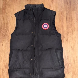 Canada Goose VÄST - MacMillan SVART - Svart dunväst från Canada Goose med klassisk logga på bröstet. Västen har hög krage, dragkedja och tryckknappar framtill samt två stora fickor. Perfekt för lager-på-lager och riktigt snygg till streetwear-stil.
