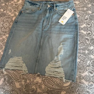 Ljusblå jeanskjol med slitningar Adpt - Trendig ljusblå jeanskjol från Adpt med råa slitningar och fransad nederkant. Klassisk femficksmodell med knapp och dragkedja framtill. Perfekt för dig som gillar en edgy och avslappnad stil.