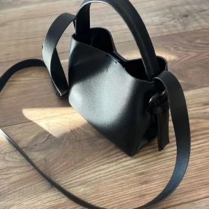 Svart stilren axelväska i skinn - Minimalistisk svart axelväska i slätt skinn med justerbar axelrem och kort handtag. Väskan har en enkel, boxig form och stängs med magnetknapp. Perfekt för dig som gillar clean och modern look.