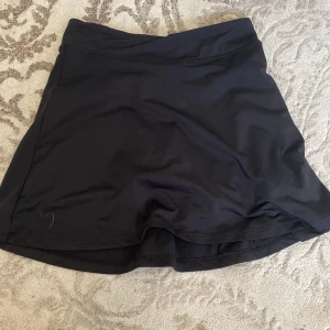 Svart sportkjol från H&M Move - Svart sportig kortkjol från H&M Move med inbyggda shorts under. Kjolen är tillverkad i stretchigt syntetmaterial och har en enkel, stilren design utan mönster eller extra detaljer. Perfekt för träning eller aktiva dagar.