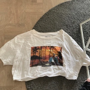 Vit croppad t-shirt med tryck Dedicated - Säljer en vit croppad t-shirt med färgglatt tryck av solnedgång, palmer och strand. T-shirten har korta ärmar och en avslappnad passform. Perfekt för dig som gillar chill vibes och snygga prints. Croppad av mig.