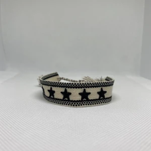 Beige och svart armband med stjärnor - Snyggt vävt armband i beige och svart med broderade svarta stjärnor runt om. Armbandet har flätade band med tofsar i ändarna för justerbar passform. Perfekt accessoar för att ge din outfit en cool touch.