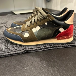Valentino Rockrunner  - Snygga Valentino Rockrunner sneakers i en cool mix av färger och material. Skorna har ett camouflage-mönster med inslag av olivgrön, marinblå och röd mocka. De har snörning och en unik nitat detalj på hälen. Perfekta för dig som vill sticka ut! 