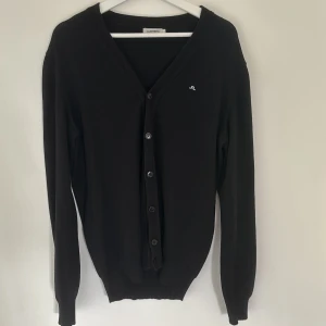 Svart kofta från J.Lindeberg - Stilren svart kofta från J.Lindeberg med v-ringning och knappar framtill. Liten broderad logga på bröstet. Långärmad och ribbade muddar vid ärmslut och nederkant. Perfekt för lager-på-lager och enkel att matcha med allt. Liten i storleken 