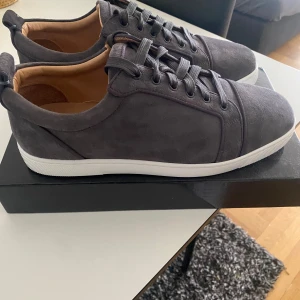 Grå mockasneakers från Louboutin - Snygga sneakers i mörkgrå mocka från Christian Louboutin med klassisk vit sula och ikonisk röd undersida. Skorna har rund tå, snörning och lyxig läderinsida. Perfekta för dig som vill ha en stilren och exklusiv look.