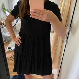 Svart kort klänning med v-ringning - Säljer en svart kortklänning med v-ringad hals och korta ärmar. Klänningen har en luftig passform med volangdetaljer i flera lager, vilket ger en avslappnad och trendig look. Perfekt för dig som gillar enkel men snygg stil.