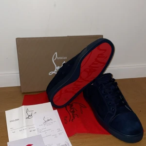 Mörkblå mocka sneakers från Louboutin - Snygga mörkblå sneakers från Christian Louboutin i lyxig mocka med klassisk röd sula. Skorna har rund tå, platt sula och snörning framtill. Perfekta för dig som vill sticka ut med exklusiv design och detaljer.
