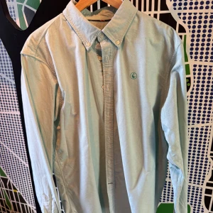 Turkos/grön skjorta från El Ganso XL - Snygg ljusblå skjorta från El Ganso i bomull. Klassisk button-down krage, broderad logga på bröstet och vita knappar. Långärmad modell med diskreta detaljer vid ärmslut och sidoslits. Perfekt för dig som gillar stilrena plagg med en twist.