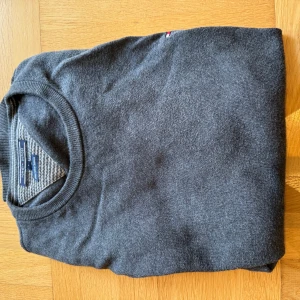 Mörkgrå tröja Tommy Hilfiger L - Mörkgrå, stilren tröja från Tommy Hilfiger i storlek L. Tillverkad i en mjuk blandning av pima bomull och kashmir. Klassisk rund halsringning, ribbade muddar och diskret logga på bröstet. Perfekt för dig som gillar en clean och tidlös look.