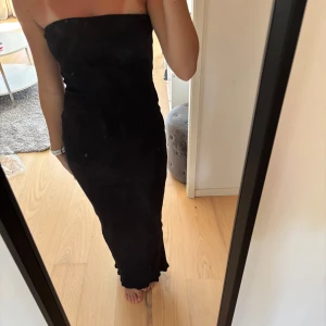 Svart axelbandslös långklänning - Säljer en svart, axelbandslös långklänning med tight passform. Klänningen har en snygg slits baktill. Materialet är ribbat och stretchigt