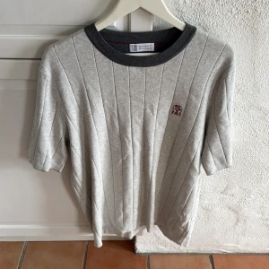 Grå randig pikétröja Brunello Cucinelli - Snygg grå pikétröja från Brunello Cucinelli med mörkgrå ribbad krage och diskret broderad logga på bröstet. Tröjan har vertikala ränder och är tillverkad i mjuk bomull. Perfekt för en clean och stilren look.