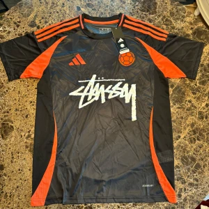Adidas x Stüssy Colombia fotbollströja M - Svart och röd fotbollströja från Adidas i samarbete med Stüssy och Colombia. Tröjan har korta ärmar, klassiska Adidas-ränder på axlarna, Stüssy-logga framtill och Colombia-tryck på ryggen. Tillverkad i lätt och ventilerande polyester med Aeroready-teknologi.