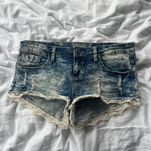 Jeansshorts med spets från Urban Surface - Snygga blå jeansshorts från Urban Surface med slitna detaljer och ljus tvätt. Shortsen har låg midja och är dekorerade med vit spets längs bensluten. Midjemått 38cm