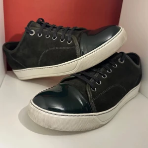 Lanvin svarta sneakers i mocka och lack - Snygga svarta sneakers från Lanvin med ovandel i mocka och lackad tå. Skorna har vita, platta sulor och svarta snören. Klassisk och stilren design med rund tå och diskreta sömmar. Perfekta för dig som gillar lyxiga detaljer och streetstyle.