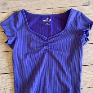 Lila ribbad topp från Hollister XS - Denna topp har använts ett fåtal gånger. Superfin lila ribbad topp från Hollister i storlek XS. Toppen har korta ärmar med lätt volangkant, v-ringning med rynkdetalj framtill och är figurnära i modellen. Perfekt att styla med jeans eller kjol för en trendig look.