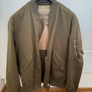 Ten C bomberjacka  - Säljer en olivgrön bomberjacka från Ten c, modell Flight Jacket A35/f. Jackan har klassisk bomberstil med ribbade muddar, dragkedja och fickor med lock framtill samt ärmficka med dragkedja. Tillverkad i bomull och polyester för en skön känsla. Nyskick , oanvänd . Köpt på Nk i Göteborg .