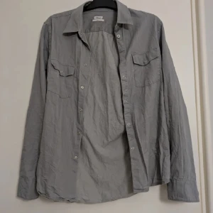Grå skjorta från Filippa K - Snygg grå skjorta från Filippa K i modellen M. Pierre Denim Shirt. Skjortan har klassisk krage, två bröstfickor med lock och knappar framtill. Långärmad och i 100% bomull. Passar till allt. 