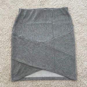 Grå ribbad kjol från Gina Tricot - Grå ribbad kortkjol från Gina Tricot med omlott-detalj framtill. Kjolen är i stretchigt material och har en snygg, figurnära passform. Perfekt att styla med en oversized hoodie eller en tight topp för en chill look.