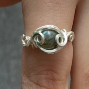 Handgjord silverring med labradorit - Unik ring i silverfärgad metall med en grågrön labradorit i mitten. Ringen har snygga tvinnade och snirkliga detaljer runt stenen som ger en bohemisk och magisk känsla. Perfekt för dig som gillar statement-smycken och vill sticka ut lite extra.