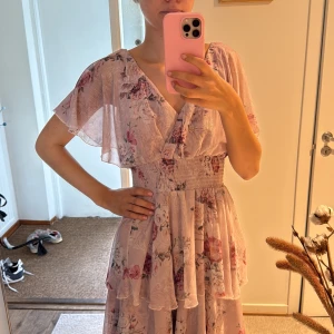 Ljusrosa blommig volangklänning - Superfin ljusrosa långklänning med blommigt mönster och flera lager volanger. Klänningen har v-ringning, korta ärmar och smock i midjan som ger en snygg passform. Perfekt för sommarens alla tillfällen!
