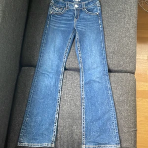 Blå bootcut jeans från Gina Tricot - Snygga blå jeans från Gina Tricot i modell bootcut med klassiska fickor och coola detaljer på bakfickorna. Jeansen har justerbar midja med knappresår och är tillverkade i jeansmaterial med kontrastsömmar.