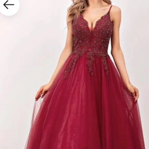 Vinröd långklänning med spets och pärlor - Magisk vinröd långklänning med djup v-ringning, tunna axelband och utsvängd kjol i tyll. Livet är dekorerat med spets och pärlor som ger en lyxig känsla. Perfekt för bal eller fest där du vill sticka ut och känna dig extra glam.