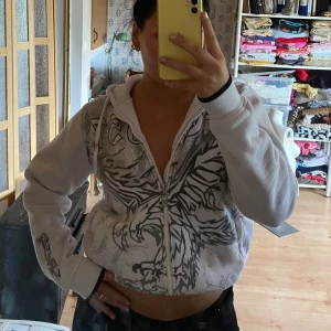 Vit hoodie med grått tribaltryck - Vit hoodie med grått tribalinspirerat örntryck över hela framsidan. Croppad modell med dragkedja, huva och snörning. Svarta detaljer vid ärmslut och tryck på ena ärmen. Perfekt för dig som gillar streetstyle och unika prints.