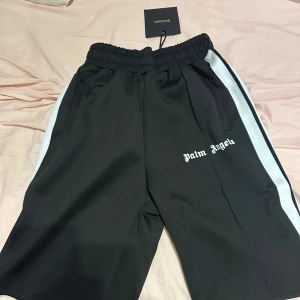 Palm Angels svarta shorts med ränder storlek S - Svarta shorts från Palm Angels med vita sidoränder och logga tryckt på vänster ben. De har elastisk midja och sportig look, perfekta för en chill streetwear-stil. Materialet känns mjukt och följsamt, och passformen är relaxed. 