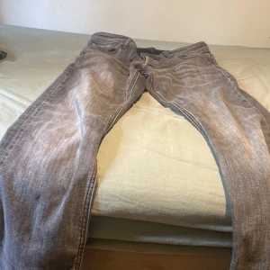 Grå jeans från Jack & Jones - Säljer ett par grå jeans från Jack & Jones med snyggt tvättad look. Klassisk femficksmodell med dragkedja och knapp i midjan. Jeansen har raka ben och normal passform, perfekta till vardagsstilen. Materialet är slitstarkt denim i bomull.
