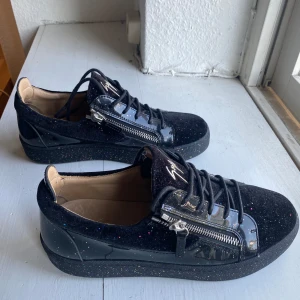 Svarta glittriga sneakers från Giuseppe Zanotti - Svarta sneakers med glittriga detaljer och signaturlogga på plösen. Skorna har snörning, dragkedjor på sidorna och en platt sula. Materialmix av mocka och lack med färgglatt glitter över hela skon. Perfekta för dig som vill sticka ut.