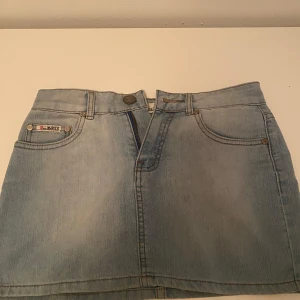 Ljusblå jeanskjol från D.realMARKS - Snygg lågmidjad ljusblå jeanskjol från D.realMARKS med klassiska fem fickor och dragkedja fram. Perfekt för dig som gillar en enkel och tidlös denimstil. Kjolen har raka sömmar och en kort passform som ger en fräsch vibe. 