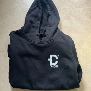 Svart hoodie från DirtCult - Svart hoodie från DirtCult med vit logga på bröstet. Klassisk huva med snörning och mjukt material som känns skönt mot huden. Perfekt för dig som gillar streetwear och enkel stil.