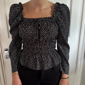 Svart blommig blus från Sisters Point - Supersöt svart blus från Sisters Point med vita små blommor, puffiga långa ärmar och smockad midja. Fyrkantig ringning med knyt framtill ger en trendig look. Perfekt för dig som gillar feminina detaljer och snygg passform.