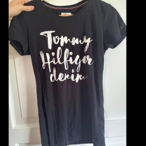 Svart t-shirt Tommy Hilfiger Denim - Svart t-shirt från Tommy Hilfiger Denim med vit tryckt text framtill. Klassisk rund halsringning, korta ärmar och liten broderad logga på ärmen. Skön bomullskvalitet och normal passform. Perfekt för en chill och stilren look.