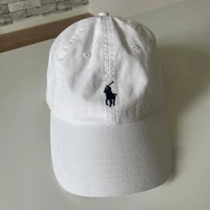 Vit keps från Polo Ralph Lauren - Snygg vit keps från Polo Ralph Lauren med klassisk broderad logga framtill och justerbart spänne bak. Kepsen är i bomull och har en stilren look med mörkblå detaljer. Perfekt accessoar för en clean och sportig stil.