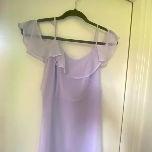 Ljuslila långklänning med volanger - Superfin ljuslila långklänning med tunna axelband och offshoulder-volanger. Klänningen har v-ringad urringning och flera lager volanger nertill för en flowy känsla. Perfekt för sommarens alla tillfällen. Materialet är tunt och lätt, troligtvis polyester.