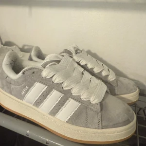 Adidas Campus grå mocka sneakers - Snygga Adidas Campus sneakers i ljusgrå mocka med klassiska vita ränder och breda vita skosnören. Skorna har en platt sula i beige och rund tå. Perfekta för dig som gillar en clean och stilren look.