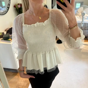 Vit blus med volang och puffärm - Supersöt vit blus med transparenta puffärmar i mesh och små prickar. Smockad överdel, volangkant upptill och nertill samt lätt utsvängd midja. 
