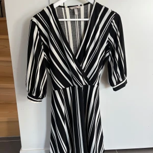 Svartvit randig omlottklänning H&M - Svartvit omlottklänning från H&M med coola diagonala och vertikala ränder. Klänningen har trekvartsärmar, v-ringning och knytband i midjan. Perfekt för dig som gillar grafiska mönster och vill sticka ut lite extra.