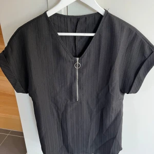 Svart randig topp med dragkedja Shein - Svart topp från Shein i storlek M med diskreta vertikala ränder och en snygg silverfärgad dragkedja framtill med ringdetalj. Toppen har korta ärmar och v-ringning, tillverkad i lätt polyester. Perfekt för en stilren och modern look.