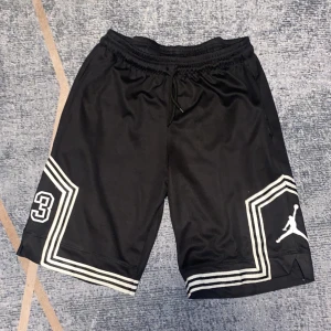 Svarta Jordan basketshorts med nr 23 - Svarta basketshorts från Jordan med vita ränder längs bensluten, tryckt Jumpman-logga och siffran 23 på ena benet. Shortsen har elastisk midja med snörning och är gjorda i ett lätt, ventilerande polyestermaterial. Perfekta för basket eller streetwear.