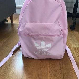 Rosa ryggsäck från Adidas - Säljer en snygg rosa ryggsäck från Adidas med det klassiska logotypen framtill. Ryggsäcken har justerbara axelremmar och en praktisk dragkedja framtill för enkel åtkomst. Perfekt för att bära dina dagliga nödvändigheter med stil. Aldrig använd.