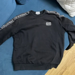 Svart sweatshirt från Emporio Armani - Snygg svart sweatshirt från Emporio Armani med EA7-logga på bröstet och längs ärmarna. Perfekt för en stilren och avslappnad look. Rund halsringning och ribbade muddar för extra komfort.