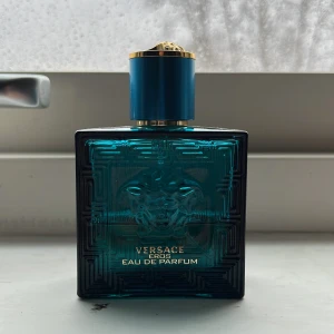 Versace Eros Eau de Parfum - Säljer en elegant Versace Eros Eau de Parfum i en stilren flaska med turkosblått glas och guldiga detaljer. Flaskan har ett klassiskt grekiskt mönster och Medusa-motiv. Perfekt för den som vill ha en doft med karaktär och stil. Det är ungefär hälften kvar alltså 25 milliliter. Luktar fantastiskt gott och är en prisvärd parfym till sommaren 