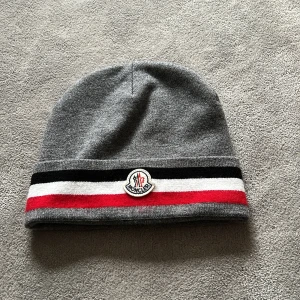 Grå mössa från Moncler - Snygg grå mössa från Moncler med röd, vit och svart randdetalj. Den har en broderad Moncler-logga framtill. Perfekt för kyliga dagar. Aldrig använd!
