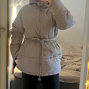 Beige vinterjacka med skärp - Varm och skön vinterjacka. Skärpet går att ta bort. Använd fåtal gånger, gott skick. Storlek 34 men något stor i modellen så passar 34-38